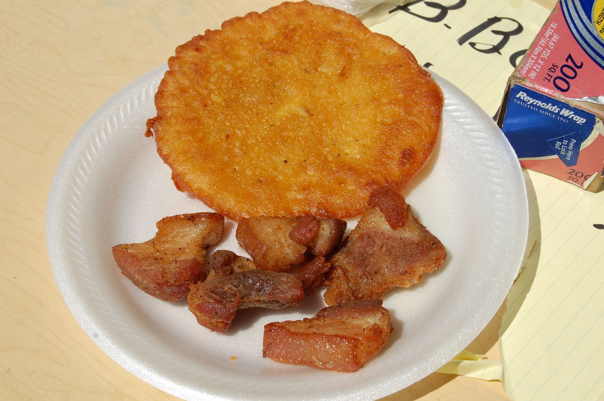 A round fried bacalaito codfish fritter next to pieces of fried pork on a paper plate
